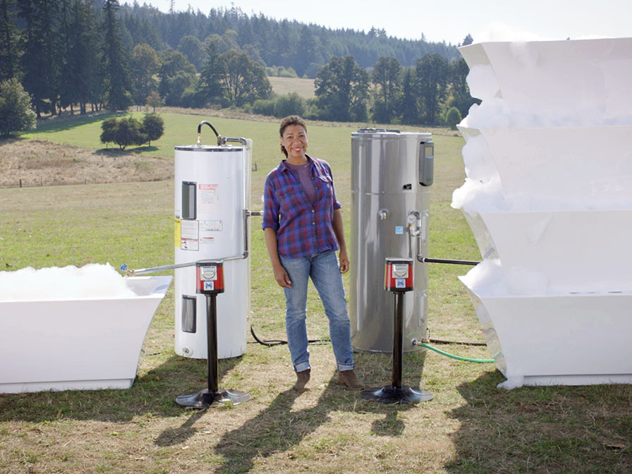 Smiling woman standing in a field. To her right is a standard water heater and a bathtub filled with soapy water. To her left is a hybrid water heater and four stacked bathtubs filled with soapy water.